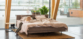Pieni poika nojailee HT Collectionin Air-sänkyyn, jossa ruskea kangasverhoilu ja pellavaiset lakanat. Kuvattu Sibeliustalossa isojen ikkunoiden edessä.