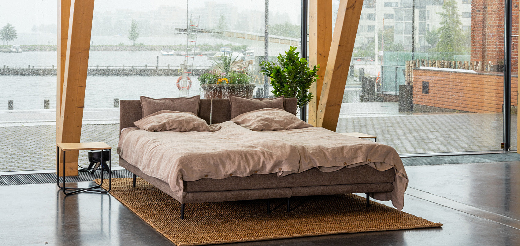 HT Collectionin Air-sänky ruskealla kangasverhoilulla ja pellavaisilla lakanoilla. Kuvattu Sibeliustalossa isojen ikkunoiden edessä, taustana Vesijärven ranta. 